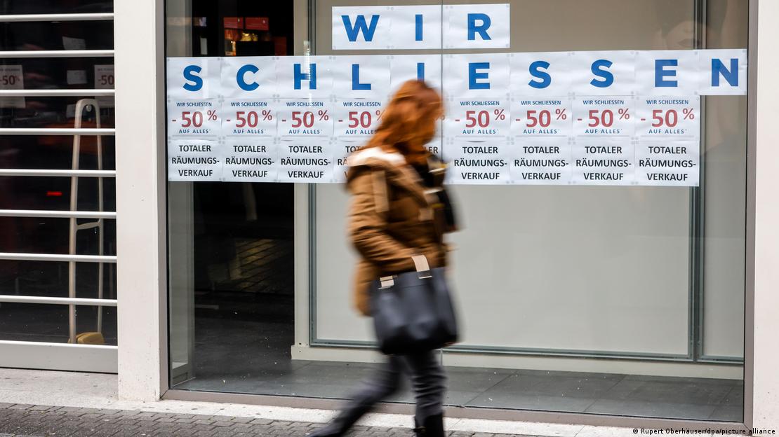 Mulher caminha em frente a uma loja com cartazes "wir schliessen", "vamos fechar", e anúncio de descontos de 50%