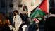 A person with dark hair, wearing a black face mask and a Palestinian keffiyeh draped over their shoulders, stands casually, looking to the left, a little in front of a statue of a seated man on plinth. A Palestinian flag is draped over the statue, and a couple more are being held on the right of the photo. There are a few other people standing nearby.
