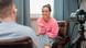 Woman in a pink sweatshirt interviewing a man in a grey shirt. A camera is trained on the young woman