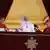 Pope Francis, a man in white clothes and a big black cross is waving from a balcony, several cardinals in red clothes and hat standing by his side.