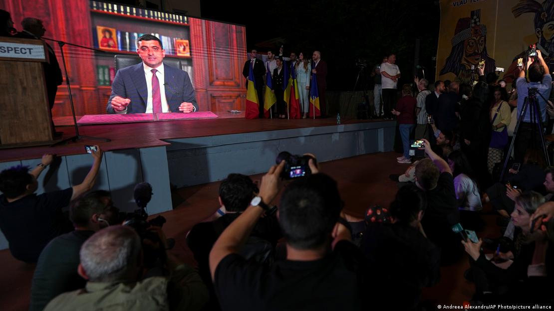 Ein Mann ist auf einem Bildschirm zu sehen. Er sitzt an einem Pult vor einem Regal, in dem Bücher stehen, die von Ikonen gesäumt sind. Vor dem Bildschirm stehen Fotografen, die das Bild aufnehmen