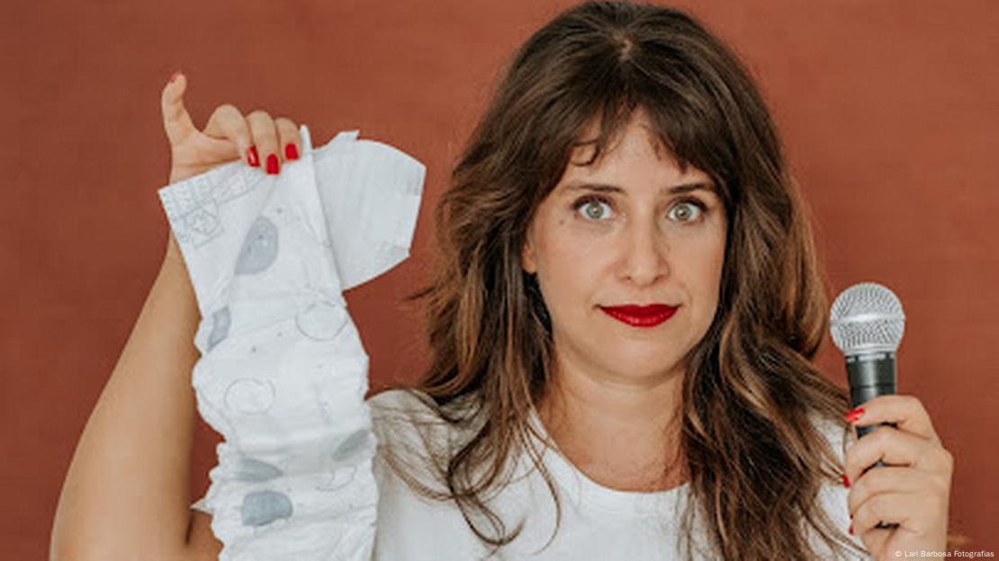 A comediante de stand-up Ana Carolina Sauwen segura uma fralda numa mão e um microfone na outra A comediante de stand-up Ana Carolina Sauwen segura uma fralda numa mão e um microfone na outra