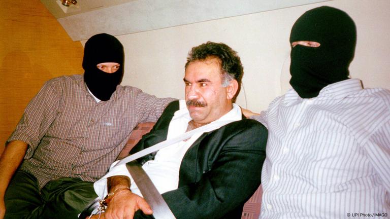 1999 yılında Kenya'da yakalanan PKK lideri Abdullah Öcalan'ın uçakla Türkiye'ye getirilirken maskeli Türk güvenlik güçleri eşliğinde elleri bağlı olarak çekilmiş fotoğrafı 1999 yılında Kenya'da yakalanan PKK lideri Abdullah Öcalan'ın uçakla Türkiye'ye getirilirken maskeli Türk güvenlik güçleri eşliğinde elleri bağlı olarak çekilmiş fotoğrafı