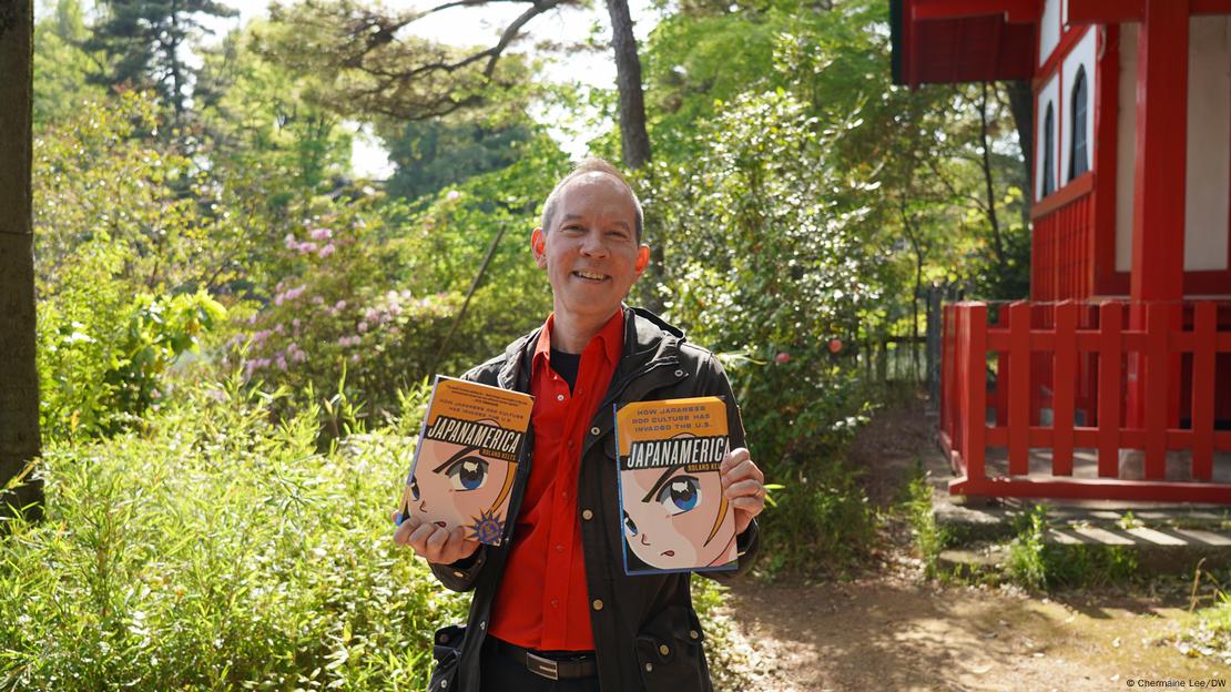 Hombre sonriendo al aire libre sosteniendo dos ejemplares del libro Japanamerica, cuya portada muestra ilustraciones de estilo anime; el fondo incluye vegetación frondosa y una estructura de arquitectura tradicional japonesa en rojo.