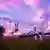 Film still from 'Twins Hinahima': in Japanese anime style, two girls standing in a field next to a bridge taking selfies at sunset.