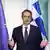 Ein Mann (Kyriakos Mitsotakis) steht an einem Mikrofon und breitet die Hände aus, während er spricht. Hinter ihm sind die griechische und die EU-Fahne zu sehen