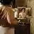 A film still from 'Sound of Falling': A woman dresses while she looks into a mirror showing different parts of her body, the room features period wallpaper.
