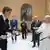 A tall man in a suit (professional tennis player Jannik Sinner) laughs with a short man dressed in white (Pope Leo XIV) as both hold tennis rackets, people and a painting can be seen in the background