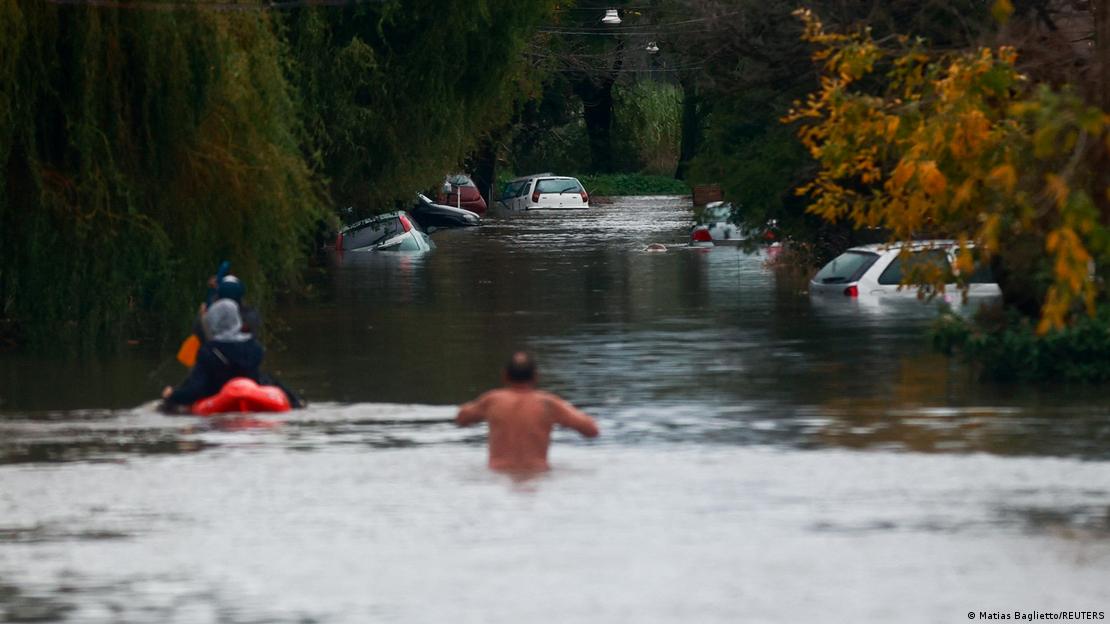 Un hombre caminando por una calle inundada de agua, en que la flotan autos a medio hundir.