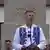 A man in a white shirt and white waistcoat embroidered with blue flowers (Peter Magyar) speaks into a microphone in front of what looks like a memorial. There are people in the background, one wearing a sash in the green, white and red of the Hungarian flag