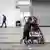 Ein kleiner Junge schiebt einen Kinderwagen in einer Geflüchtetenunterkunft. In dem Kinderwagen sitzt ein Mädchen. Beide Kinder sind von hinten zu sehen.