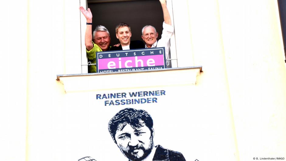 Fasbinderov portert a gore su vlasnici hotela i gradonačelnik Minhena. „Dojče ajhe“ (Nemački hrast) važi za jedno od najstarijih mesta okupljanja homoseksualaca u Minhenu. Na fasadi se već nalazi mozaik portret Fredija Merjkuruja koji je tu živeo nekoliko godina. U periodu od 1921. do 1923. navodno je i Adolf Hitler rado posećivao to mesto – kako se pričalo, zbog baletana, a i sedište NSDAP je bilo u blizini.