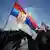 The sun shines through a large Serbian flag being carried during a protest. People, some carrying flags or signs, walk along a road, Belgrade, Serbia, June 1, 2025