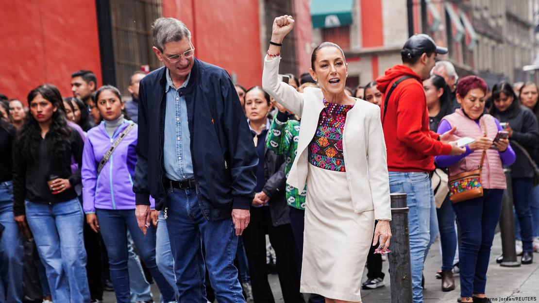 Claudia Sheinbaum com punho erguido, acompanhada de homem, enquanto pessoas observam