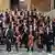 A large stringed orchestra stand on the steps of a building and poses