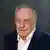 Frederick Forsyth wearing a white shirt and dark blue jacket looks at the camera