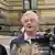 A gray-haired man in a pink shirt and blue jacket (Compact magazine editor-in-chief Jürgen Elsässer) seen speaking to reporters with microphones and cameras in front of the stone facade of the Federal Administrative Court in Leipzig, Germany on June 10, 2025