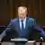 Ein Mann in einem Anzug mit blauer Krawatte, Donald Tusk, steht an einem Redepult vor einer holzverkleideten Wand