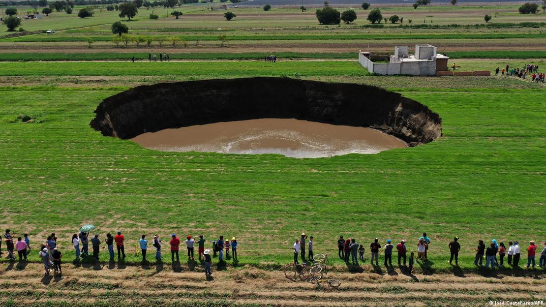 Imagem de drone mostra pessoas olhando para grande cratera em meio a plantação