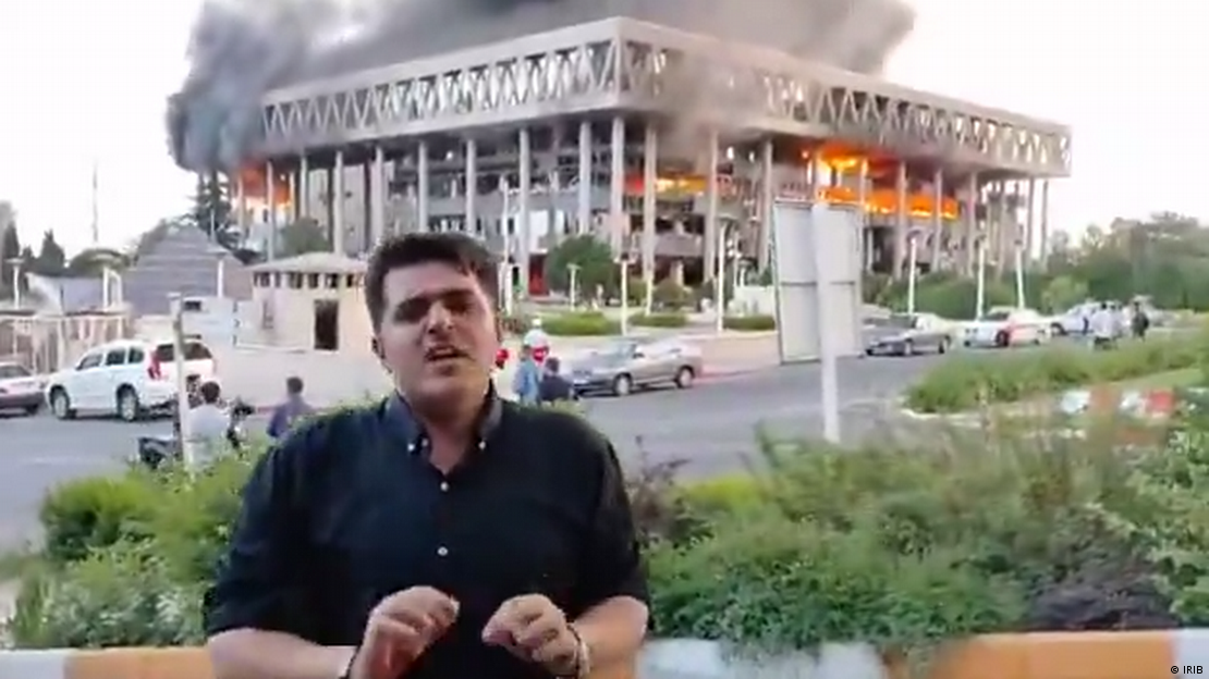 Imagem em baixa resolução mostra jornalista em frente à sede da Irib, a TV estatal iraniana, com partes do edifício em chamas