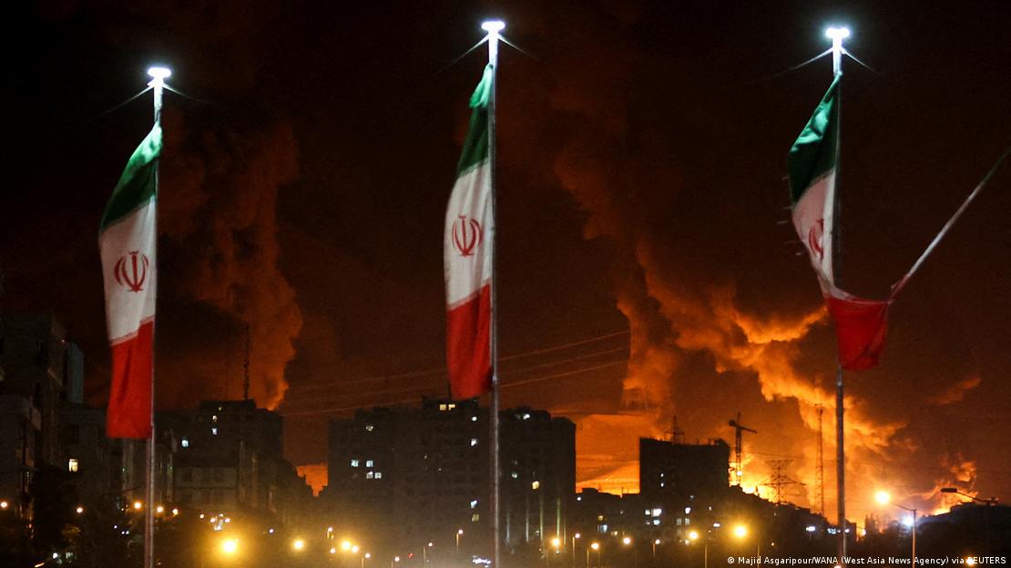 İran bayraklarının gerisinde, İsrail'in Şehran petrol deposunu hedef aldığı saldırı sonrasındaki patlamalar görülüyor.