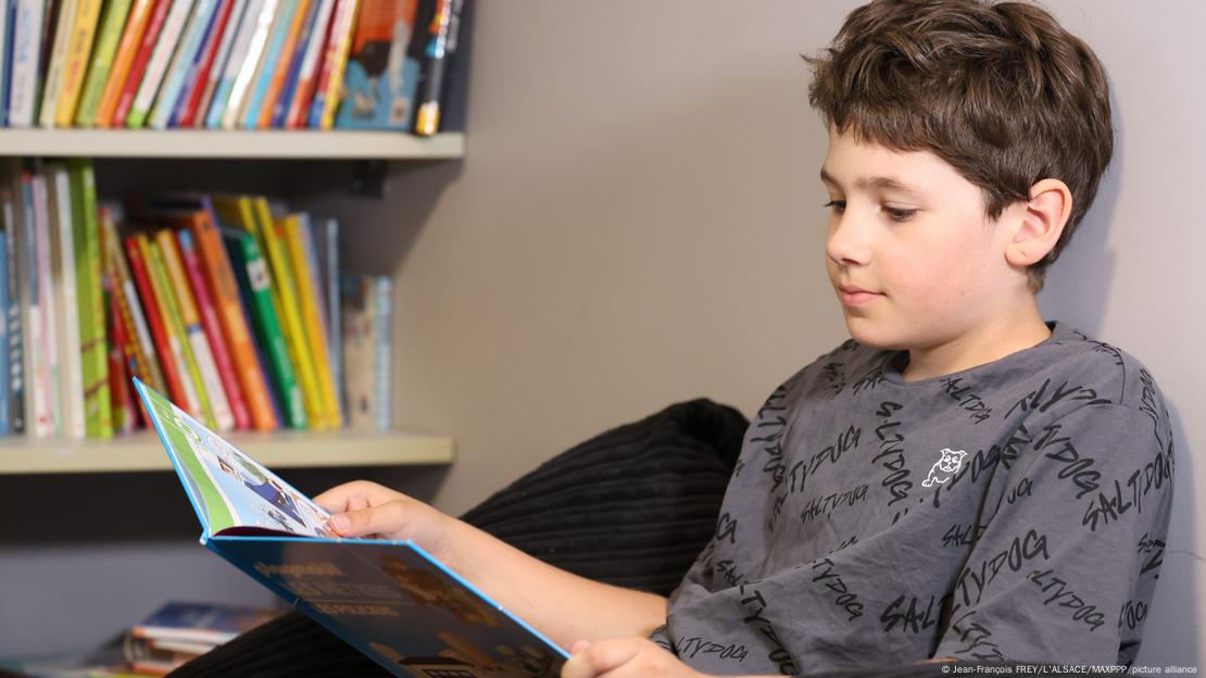 Un niño de 10 años lee un libro delante de una estantería.