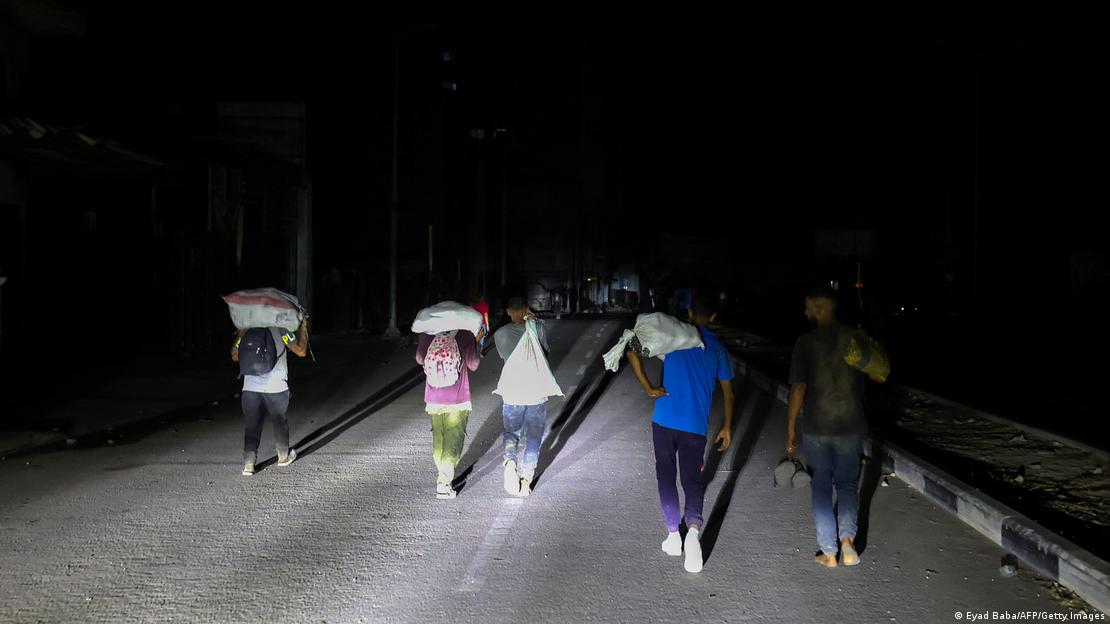 In der Dunkelheit sind auf einer Straßen einige Menschen von hinten zu sehen, die Säcke auf den Schultern oder dem Kopf tragen