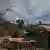 Firefighter works to put out fire near houses by hill with trees; the hill is covered in smoke