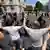 Civilians hold up their hands as they face a line of police officers in riot gear at an intersection. Other civilians and police officers face off in a similar way on the other side of the intersection in the background. Belgrade, Serbia, Monday, June 30, 2025