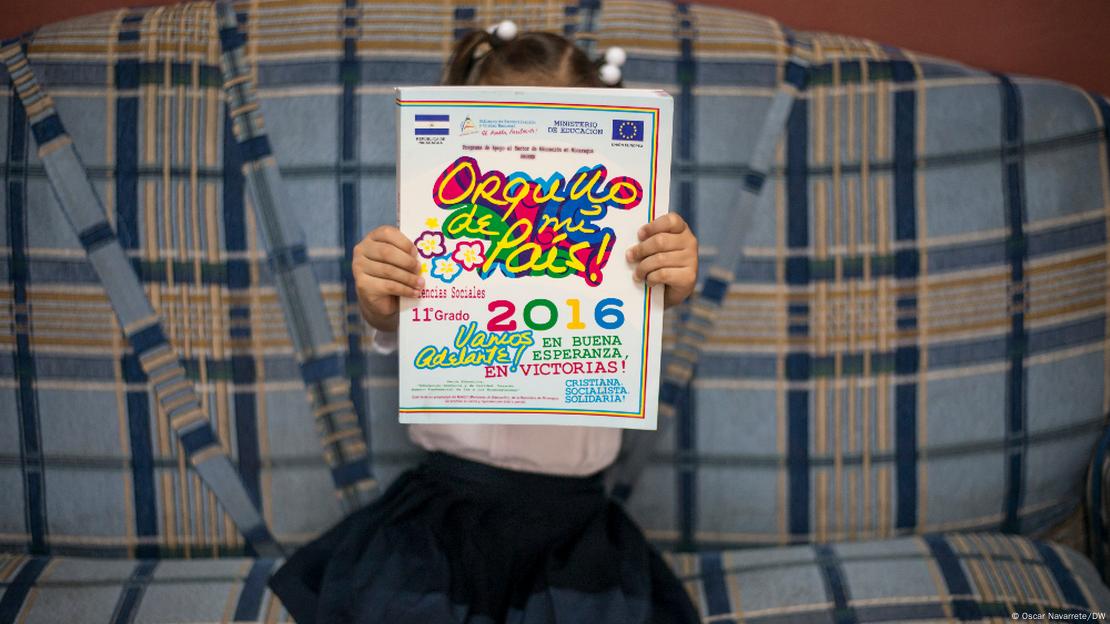 Una niña con uniforme de escuela pública muestra uno de los textos escolares con el eslogan del gobierno: "Vamos adelante, en buena esperanza y en victorias". Una niña con uniforme de escuela pública muestra uno de los textos escolares con el eslogan del gobierno: "Vamos adelante, en buena esperanza y en victorias".
