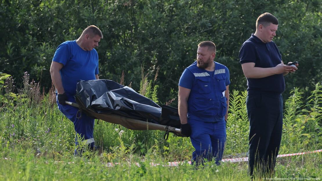 Двајца мажи во сини маици и панталони на носилка носат тело во црна вреќа. Покрај нив, на трева, стои маж облечен во црно, кој гледа во својот телефон. Двајца мажи во сини маици и панталони на носилка носат тело во црна вреќа. Покрај нив, на трева, стои маж облечен во црно, кој гледа во својот телефон.