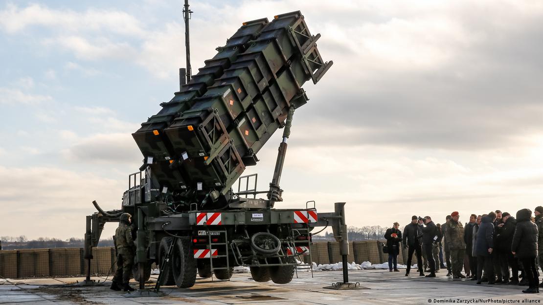 Ein Patriot-Raketenabwehrsystem vor einem bedeckten Himmel. Daneben stehen uniformierte Männer