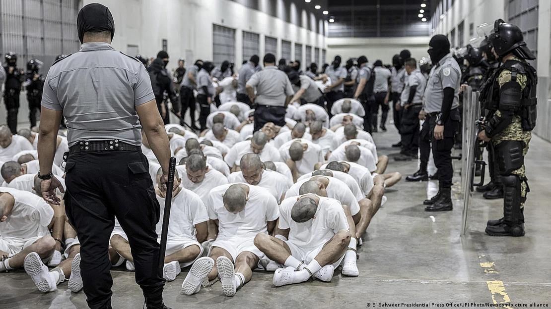 Sentados en el suelo, con la cabeza gacha y las manos esposadas a la espalda, vestidos de blanco y rodeados de agentes de seguridad en uniformes grises y negros, presuntos integrantes del Tren de Aragua y la MS13 en la cárcel CECOT, en El Salvador. Sentados en el suelo, con la cabeza gacha y las manos esposadas a la espalda, vestidos de blanco y rodeados de agentes de seguridad en uniformes grises y negros, presuntos integrantes del Tren de Aragua y la MS13 en la cárcel CECOT, en El Salvador.