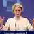 Portrait von EU-Kommissionspräsidentin Ursula von der Leyen. Sie trägt einen beigefarbenen Blazer und hält beide Hände nach vorne.