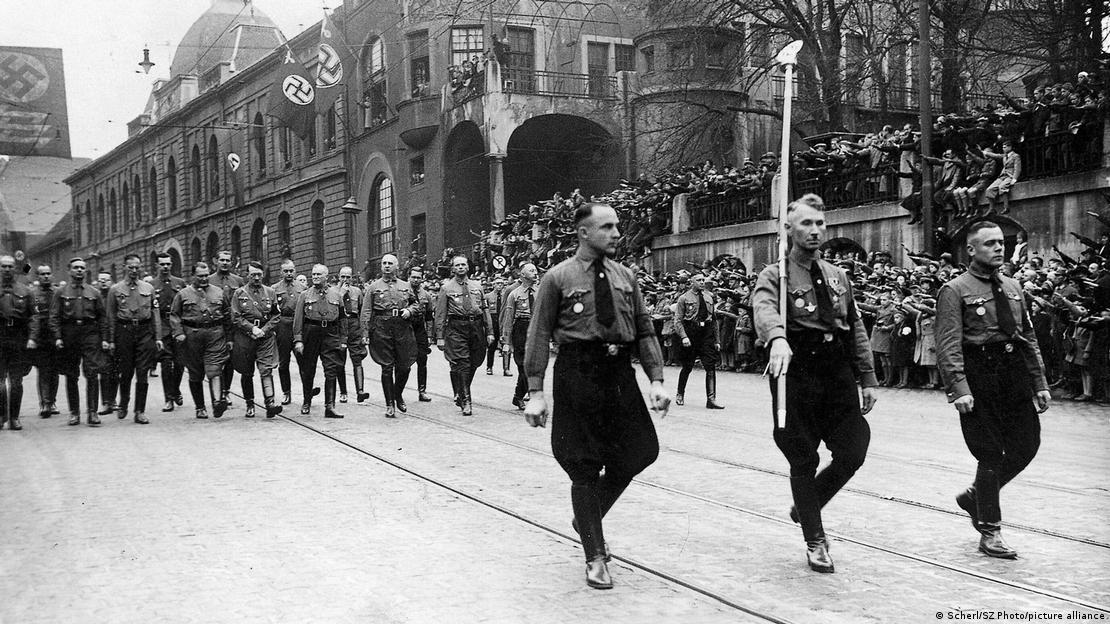 Nazis march on the street with their symbols visible on banners in the background