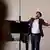 Joven canta con brazos abiertos, de fondo, un piano