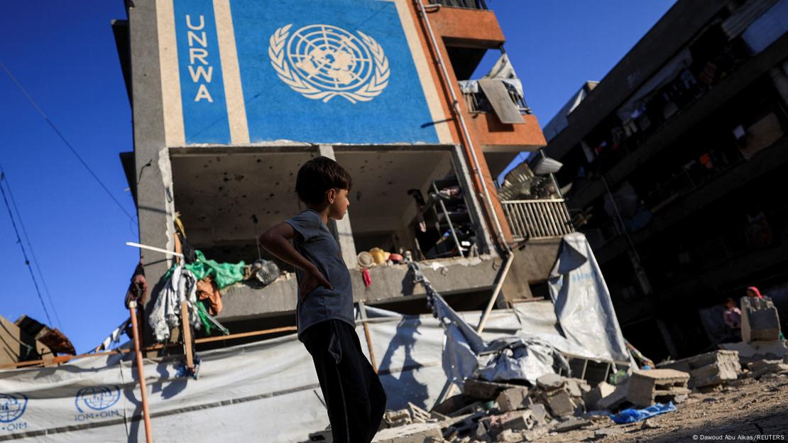 Criança ao lado de prédio semidestruído em Gaza com o símbolo da UNRWA pintado na lateral