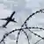 An airplane is seen in the sky, in the foreground barbed wire