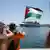 Eine Frau in orangenem Shirt schwenkt eine palästinensische Flagge. Im Hintergrund ist ein Kreuzfahrtschiff zu sehen