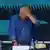 Ein Mann (Viktor Orban) in einem blauen Hemd mit offenem Hemdkragen steht mit gesenktem Kopf hinter einem Tisch und hält sich die Hand an die Stirn