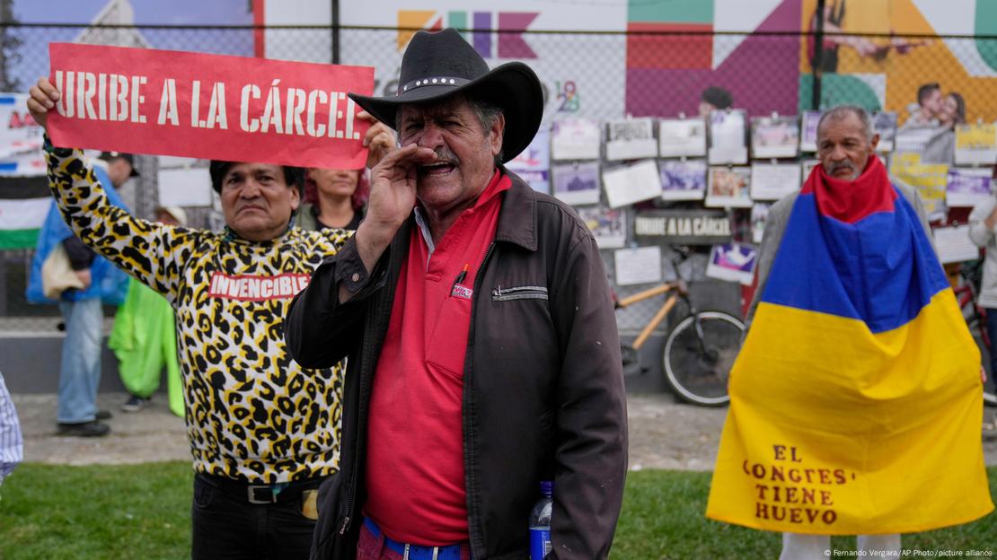 Hombre con sombrero. Detrás un hombre se cubre con bandera de Colombia. Una mujer lleva una pancarta: "Uribe a la cárcel". Hombre con sombrero. Detrás un hombre se cubre con bandera de Colombia. Una mujer lleva una pancarta: "Uribe a la cárcel".