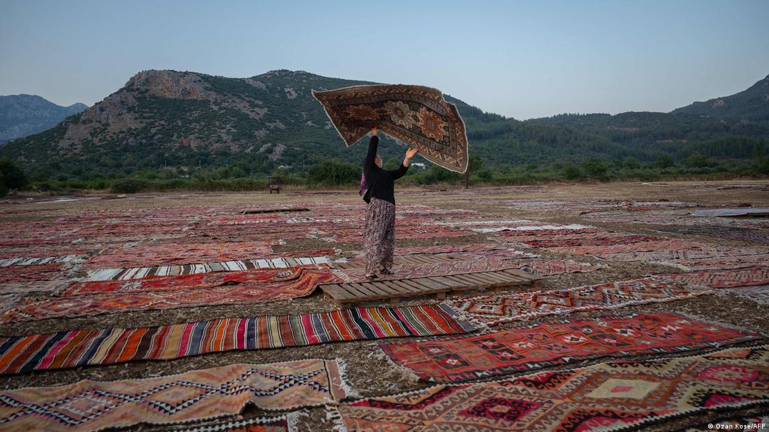 Köylü bir kadın, havalanması için güneşin altına serilen el dokuma halıları arasında bir halıyı silkelerken  