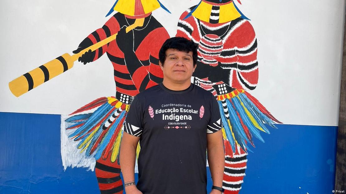 O professor indígena Mariel Kujiboekureu em frente a um mural colorido mostrando homens indígenas