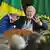 Lula da Silva habla con manos en alto, con 2 micrófonos parado tras una tarima. lo escuchan varios hombres, de fondo, bandera de Brasil.