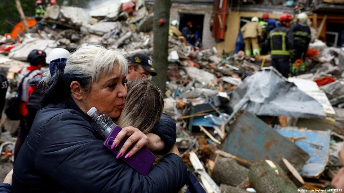 mujer abraza a otra, atrás destrucción total de viviendas y bomberos trabajando.