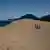 Three people shown from afar walking across a sand dune with sea visible in the background (July 28, 2025)