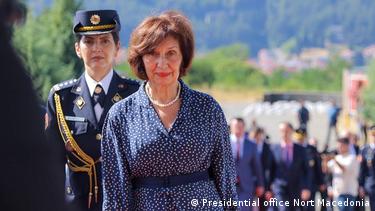 Македонската претседателка Гордана Сиљановска-Давкова облеќена во свечен темно син фустан со бели точки и ланче од бисери на вратот оди во преден план. Зад неа е жена во униформа