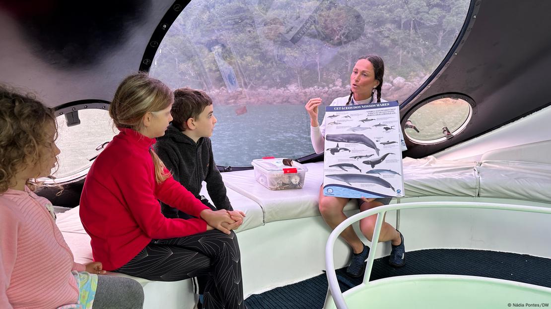 Uma mulher falando com crianças em um barco com grandes janelas redondas no mar