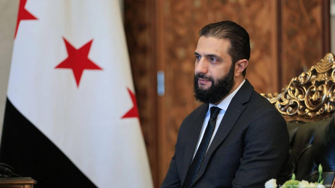 A imagem mostra o atual presidente interino da Síria, Ahmad al-Sharaa, com parte da bandeira síria ao fundo. Ahmad tem barba escura e grande, veste terno e gravata escuros, e camisa branca. A imagem mostra o atual presidente interino da Síria, Ahmad al-Sharaa, com parte da bandeira síria ao fundo. Ahmad tem barba escura e grande, veste terno e gravata escuros, e camisa branca.
