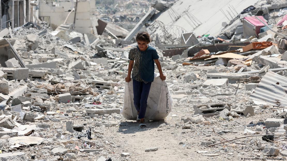 Etrafı bina yıkıntıları ile çevrelenmiş bir kız çocuğu ardında bir çuvalı sürükleyerek ilerliyor